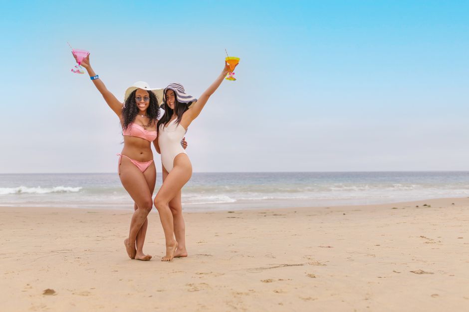 Two women enjoying a sunny day at the beach, raising colorful drinks with smiles and sun hats.