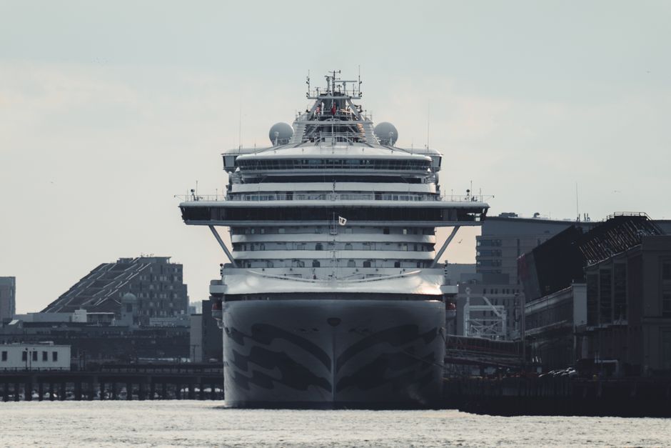A massive cruise ship docked at an urban harbor, amidst tall buildings and clear skies.