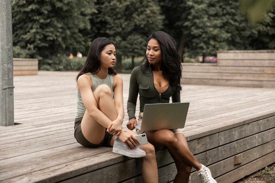 Stylish young multiethnic girlfriends with long dark hair in casual clothes resting in green park with laptop and gossiping