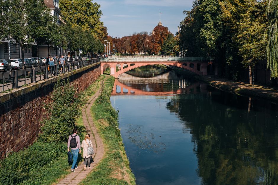 Wide calm river under stone bridge near green park and walkway in modern city