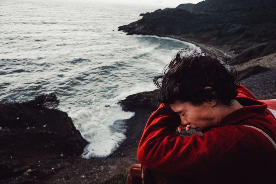 A woman in a red jacket sits by a rocky ocean shore, eyes closed, enjoying the coastal breeze.