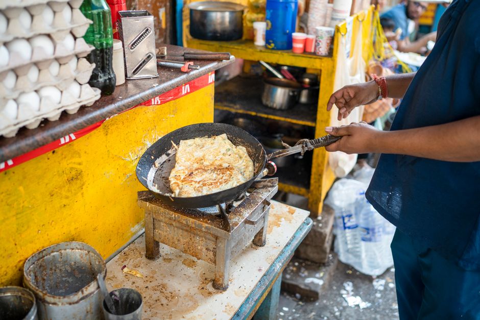 Street vendor prepares omelette in frying pan at bustling market stall with fresh eggs and ingredients.