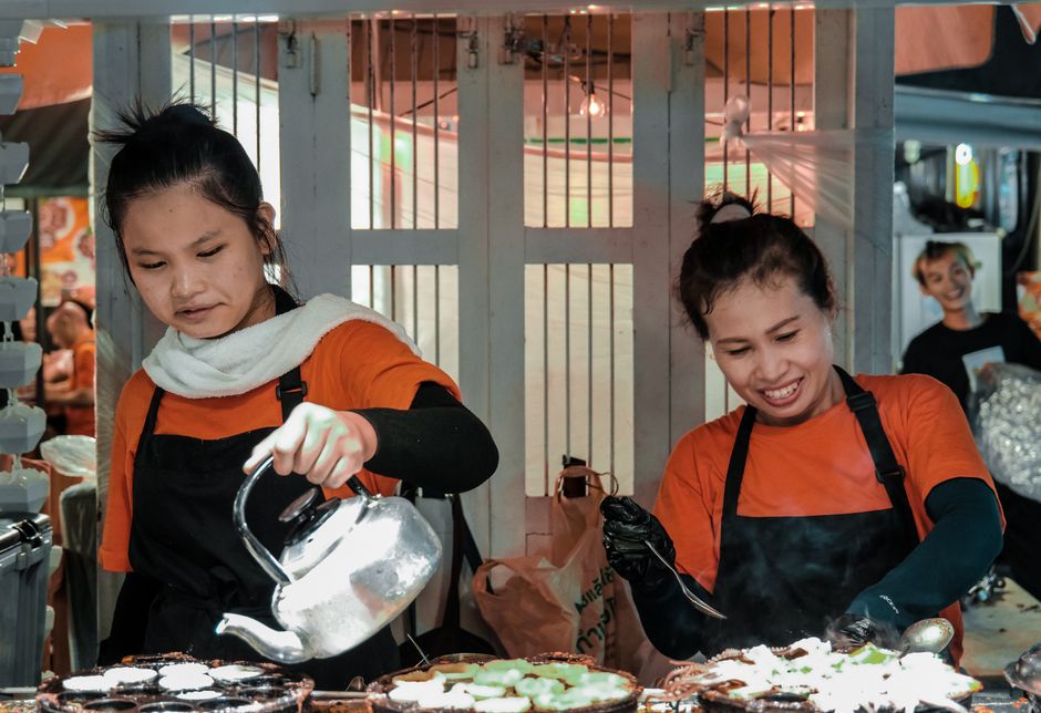 Two women prepare food with joy at a bustling Bangkok street market.