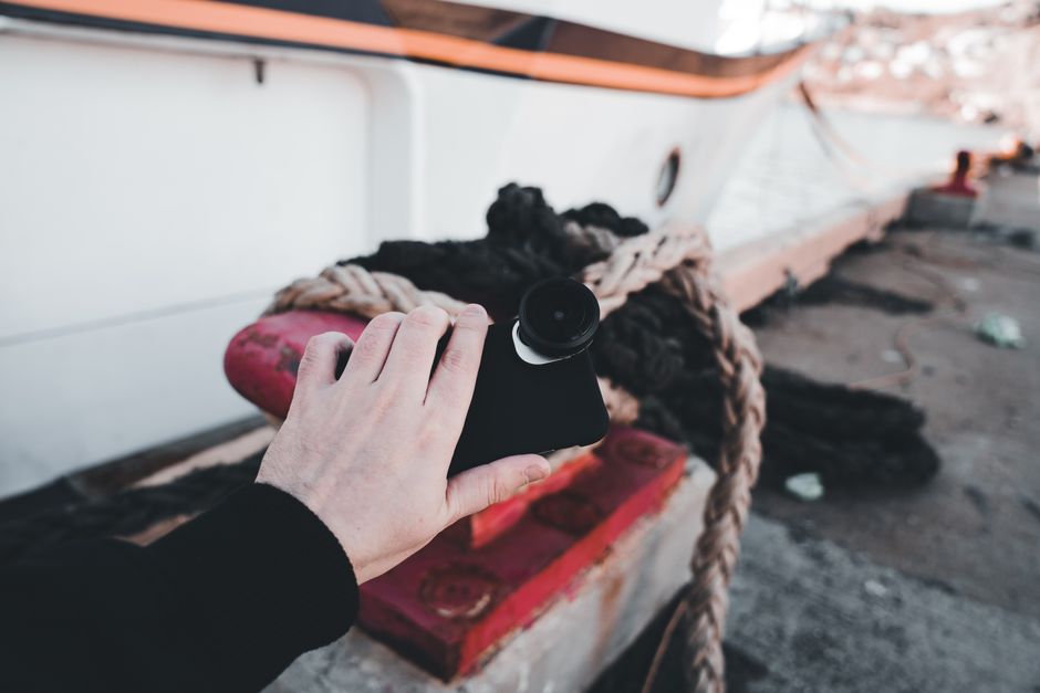 A hand holding a smartphone with fisheye lens by the harbor, showcasing technology and travel.