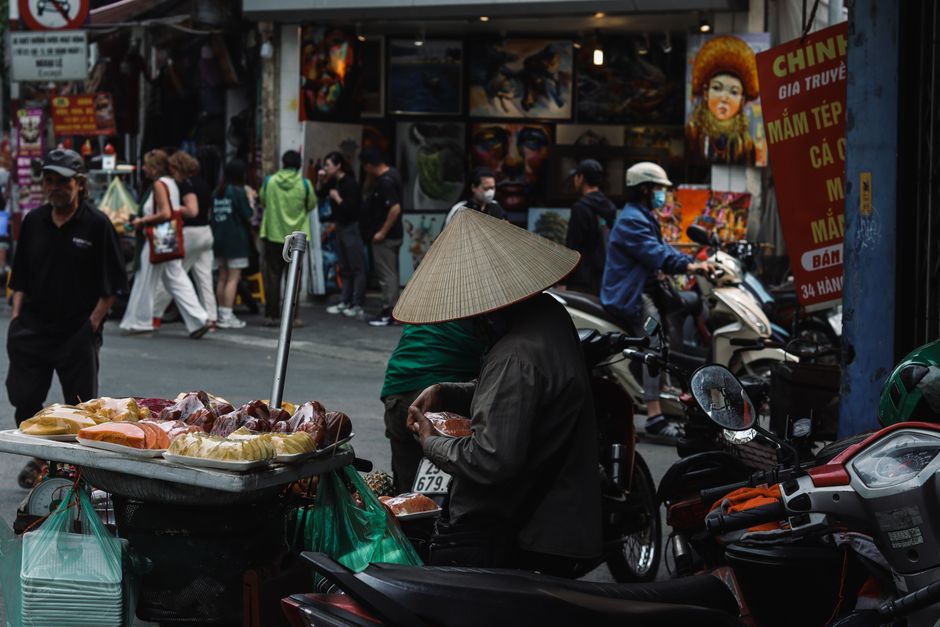 Street vendor with traditional conical hat selling fruits at a busy Vietnamese market.