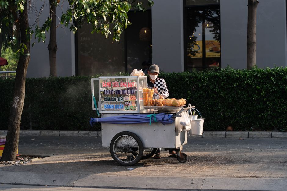 A street vendor with a cart selling bánh tiêu and other pastries on a sunny day.