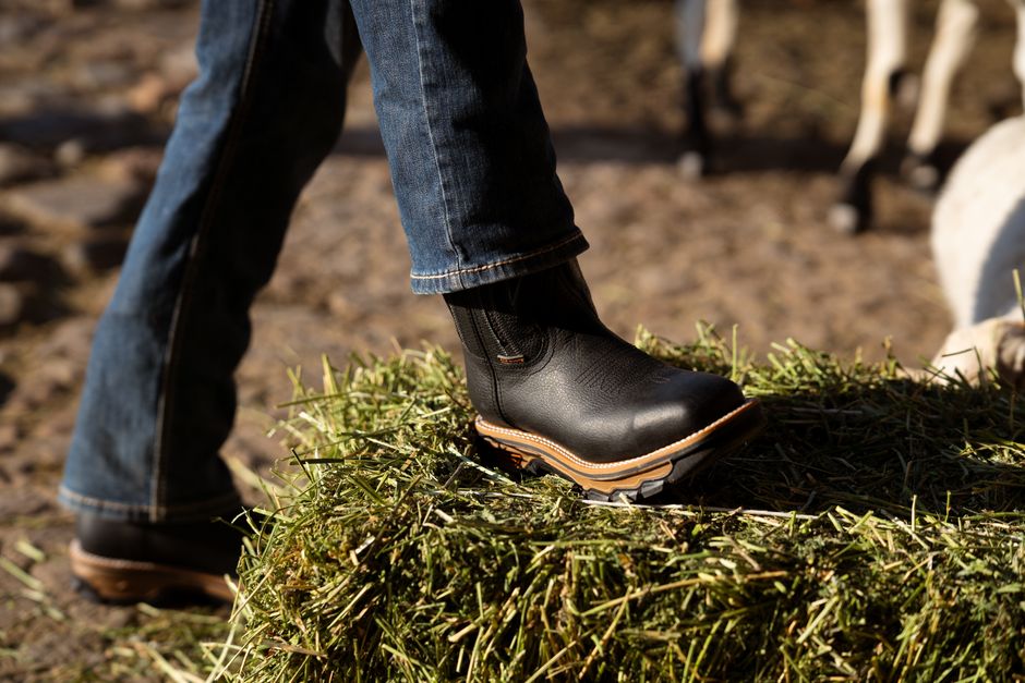 A close-up shot of a black leather boot stepping on a bale of hay outdoors, suggesting a rural lifestyle.